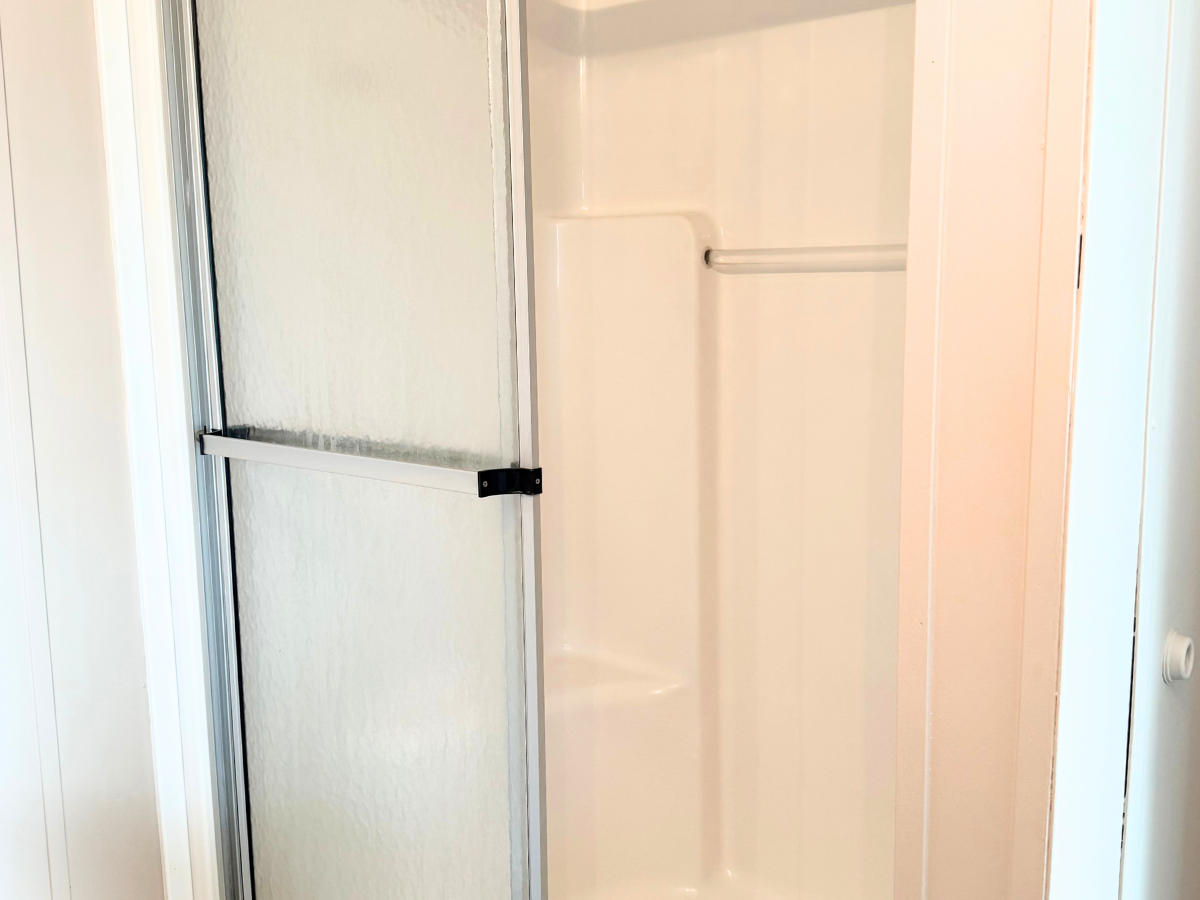 A small shower with a sliding glass door, white interior, built-in corner shelves, and a metal towel bar inside.