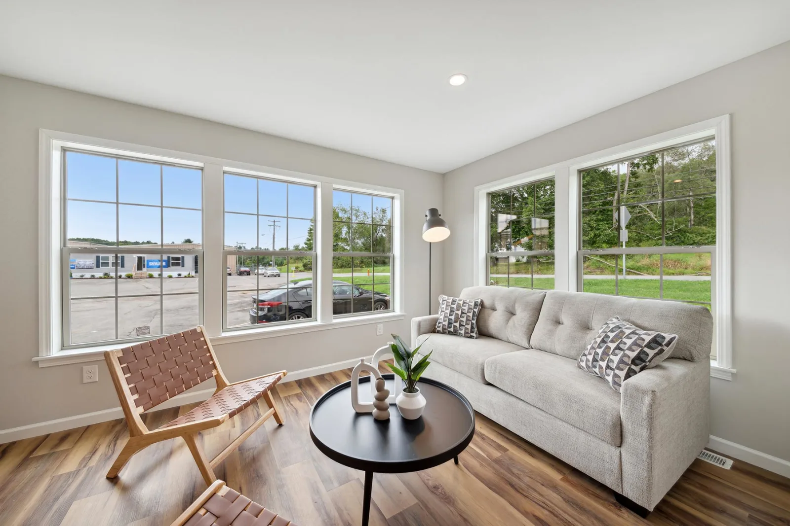 A bright living room with large windows, a gray sofa, two woven chairs, a round black coffee table, and wood flooring overlooking a parking lot and greenery outside.