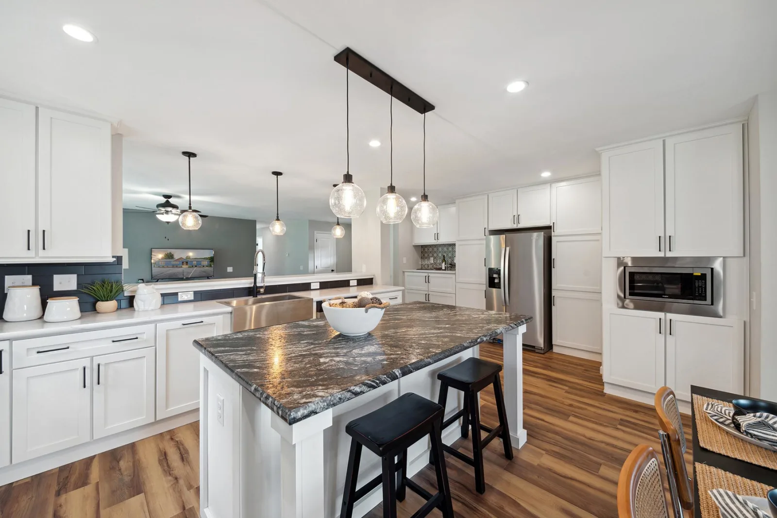 Modern kitchen with white cabinets, stainless steel appliances, marble island countertop, pendant lights, and two black stools on a wood floor.