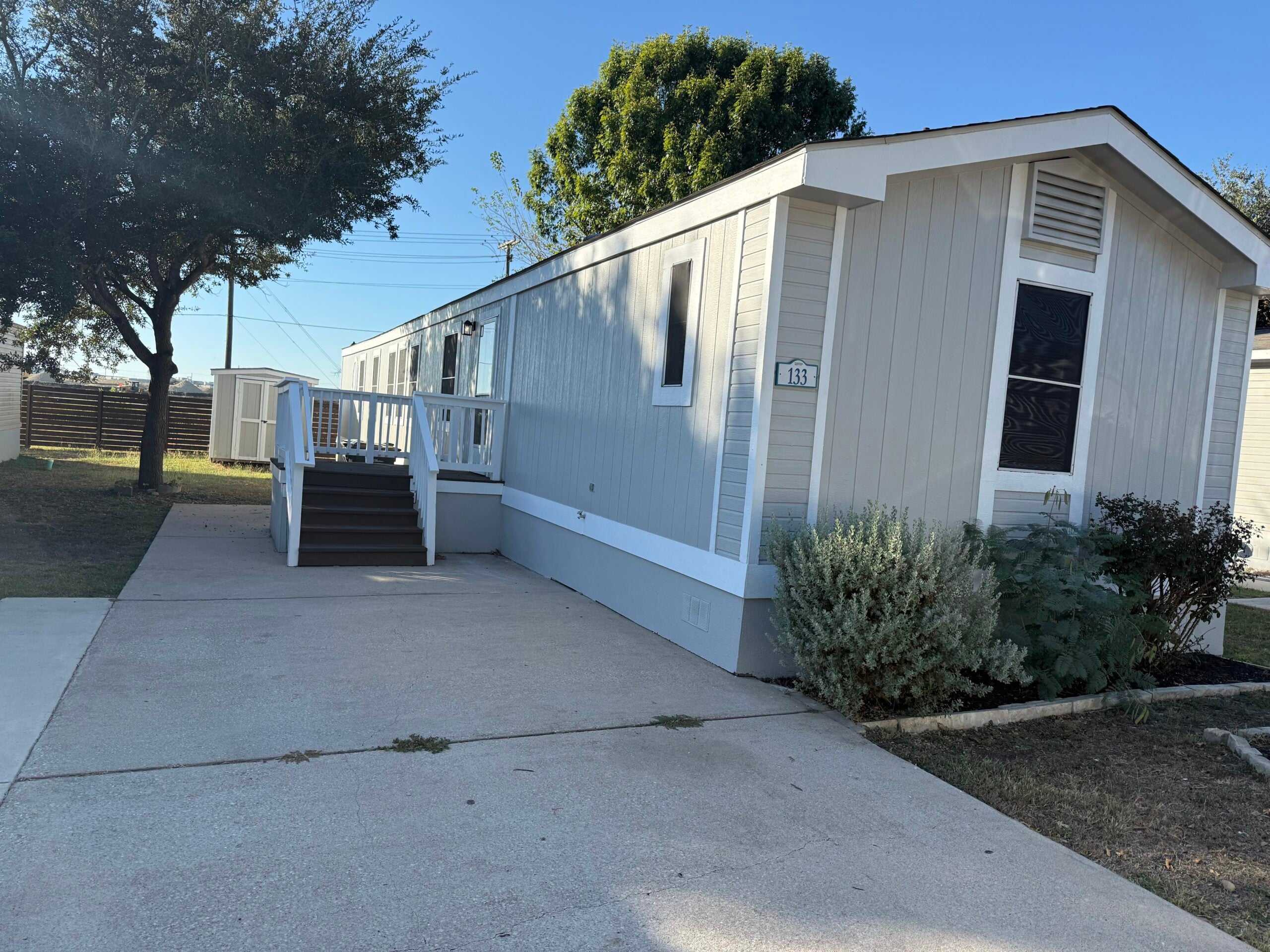 A light gray mobile home with white trim, ramp and stairs, sits on a concrete driveway with shrubs and trees nearby under a clear blue sky.