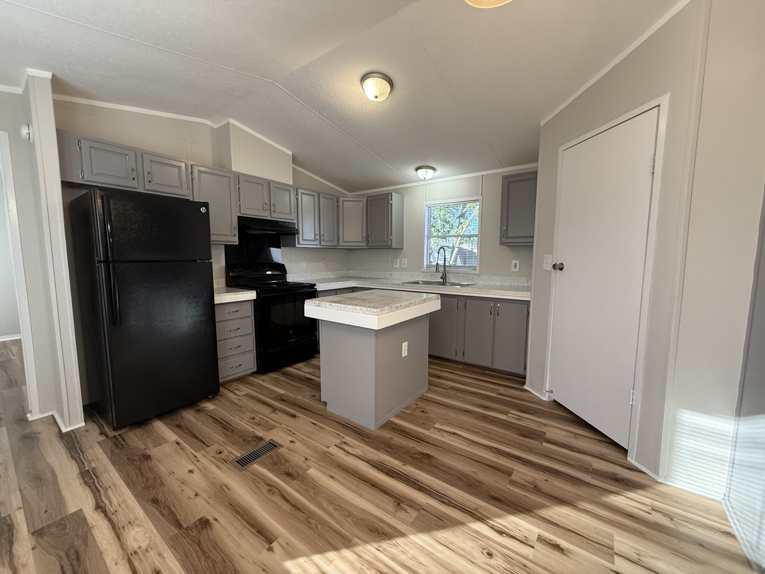 Modern kitchen with gray cabinets, black appliances, a small center island, wood-look flooring, and a window above the sink.