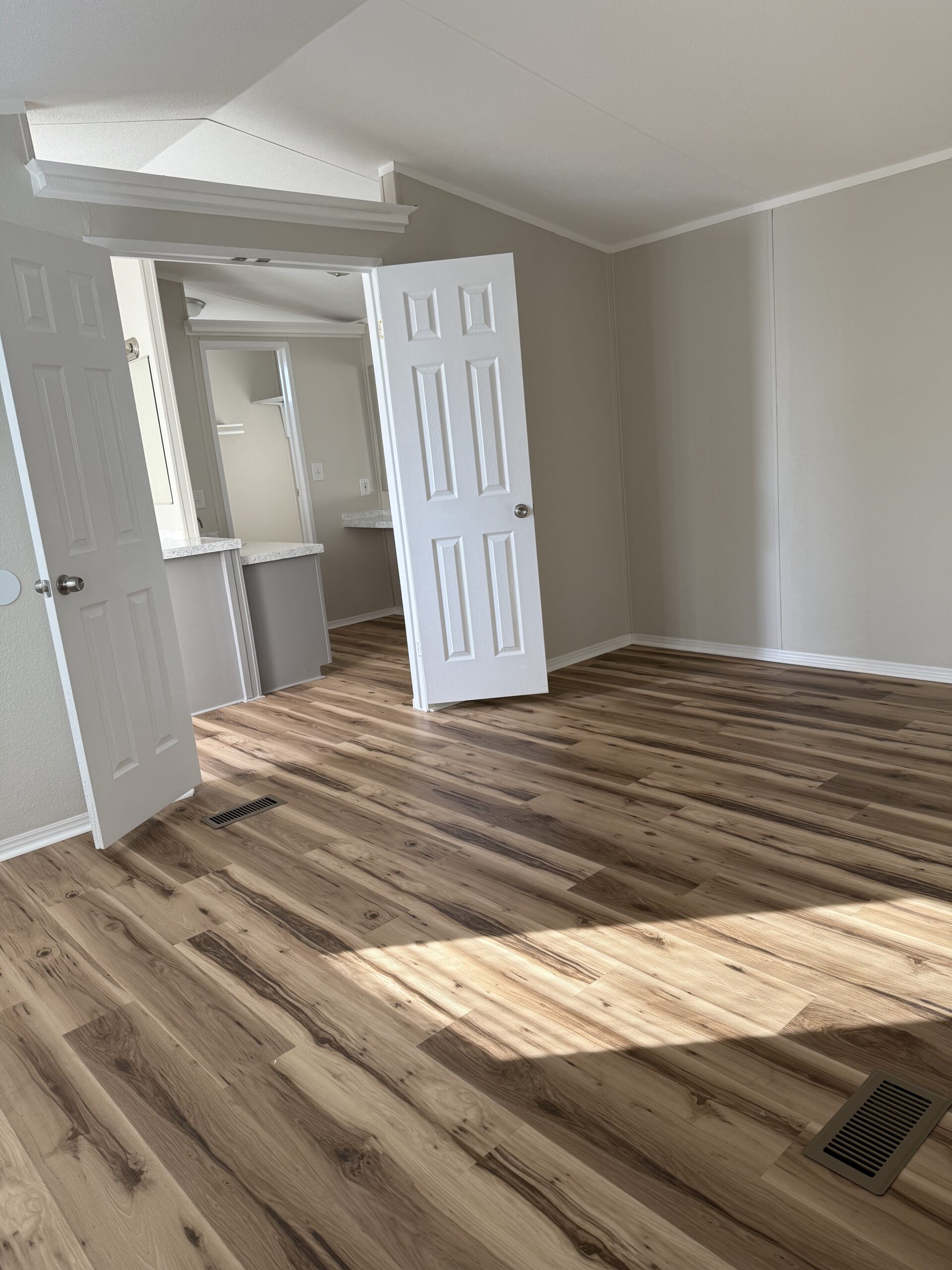 Empty room with light wood flooring, beige walls, a white ceiling, and two open doors leading to a bathroom and a closet. Sunlight is casting shadows on the floor.