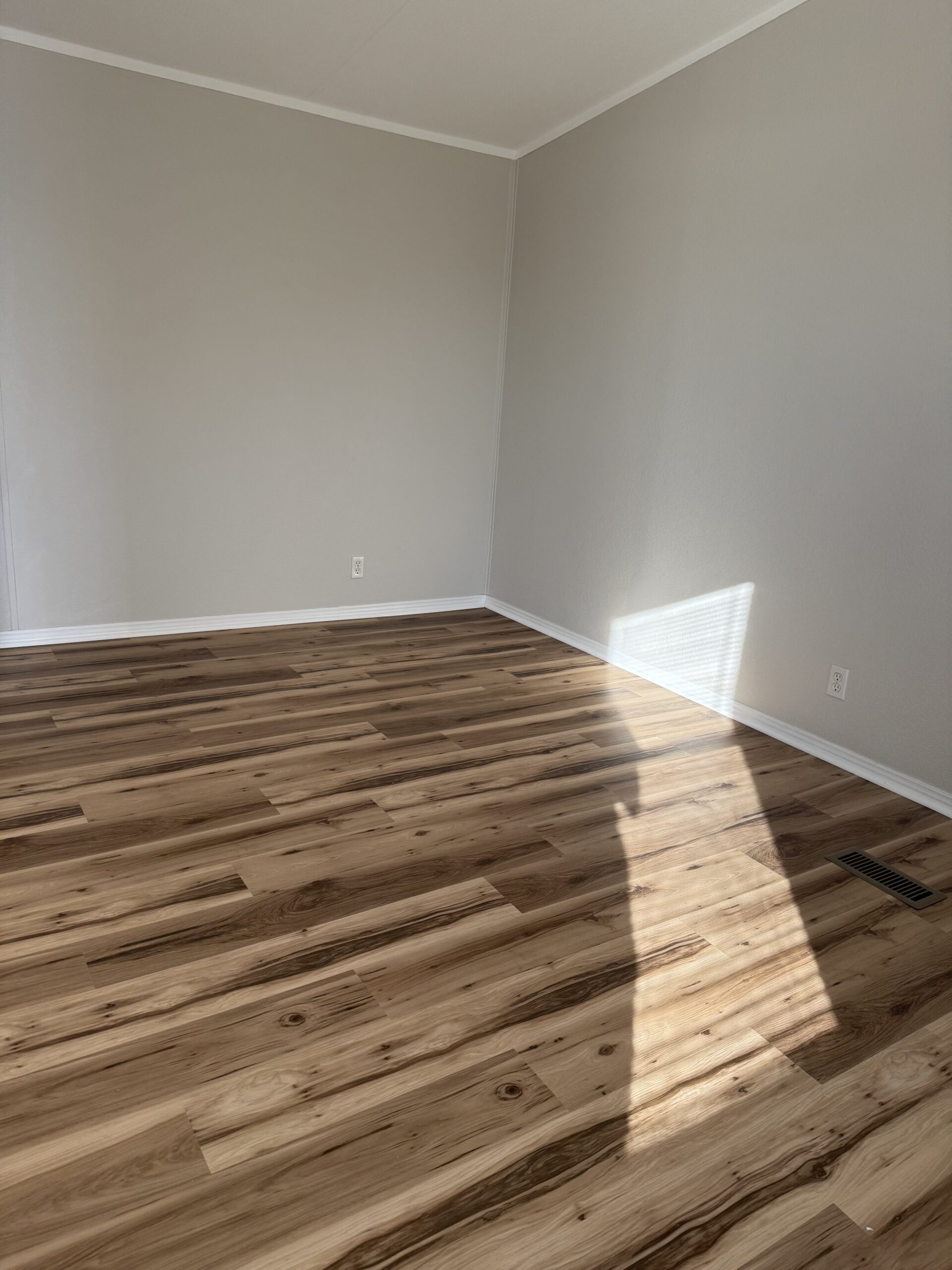 Empty room with light gray walls, white trim, and natural wood flooring. Sunlight streams through a window, casting shadows on the floor.