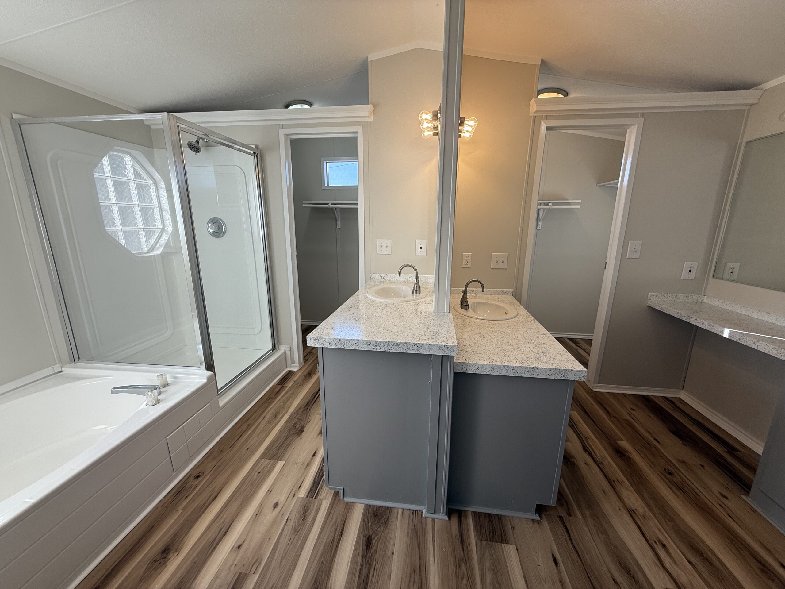 Modern bathroom with double sinks, glass shower, soaking tub, wood-look flooring, and walk-in closet visible in the background.