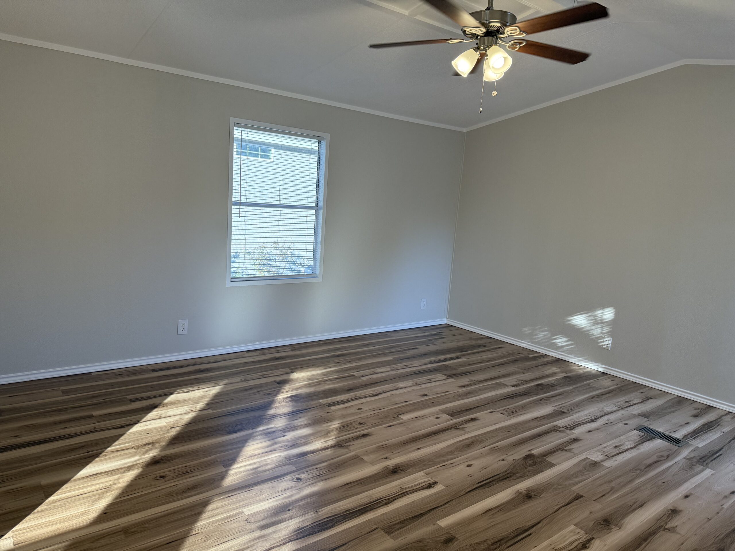 Empty room with light-colored walls, a window with blinds, wooden flooring, and a ceiling fan with lights. Sunlight is casting shadows on the floor.