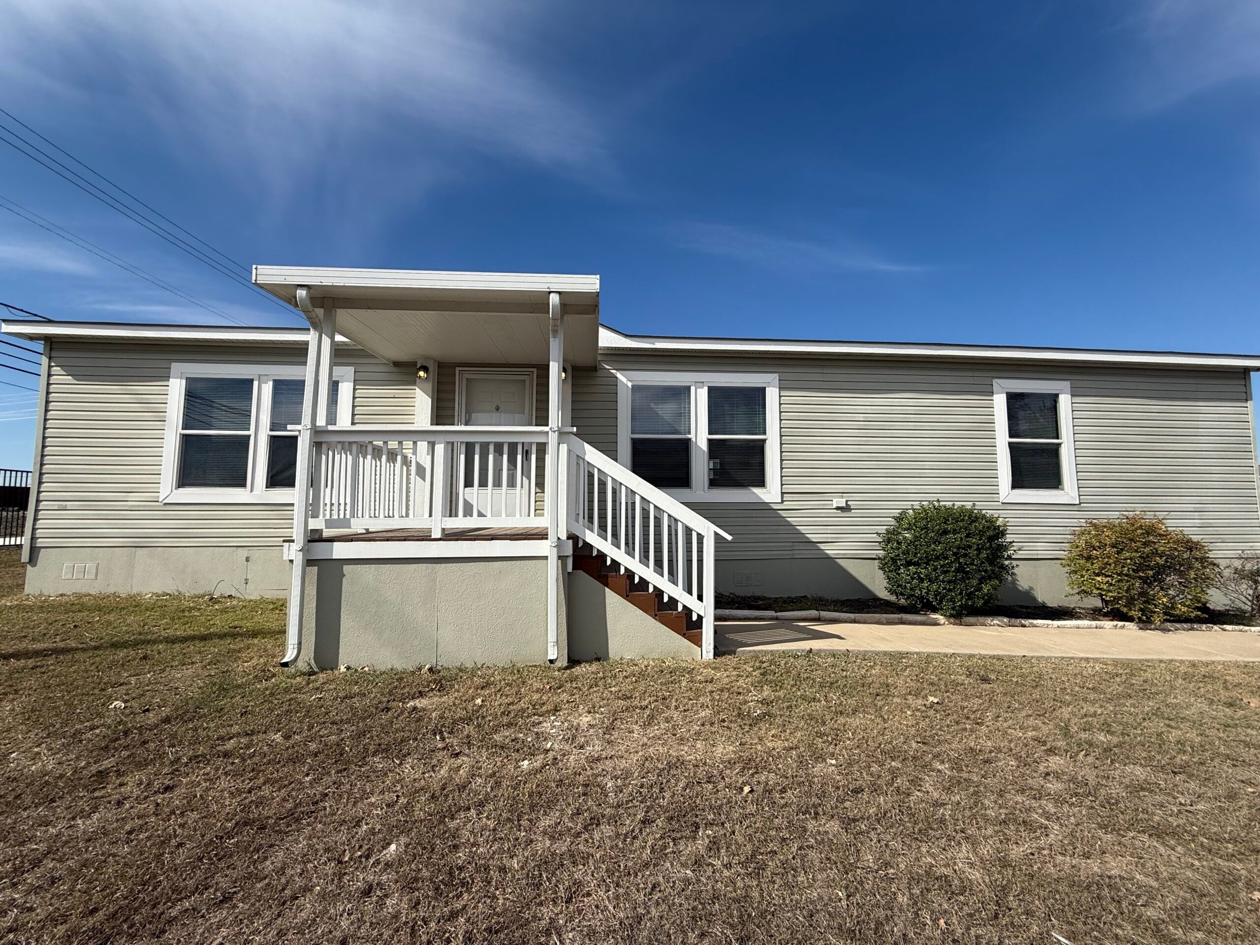 A single-story beige manufactured home with a small front porch, white railing, and stairs, set on a dry grassy yard under a blue sky.
