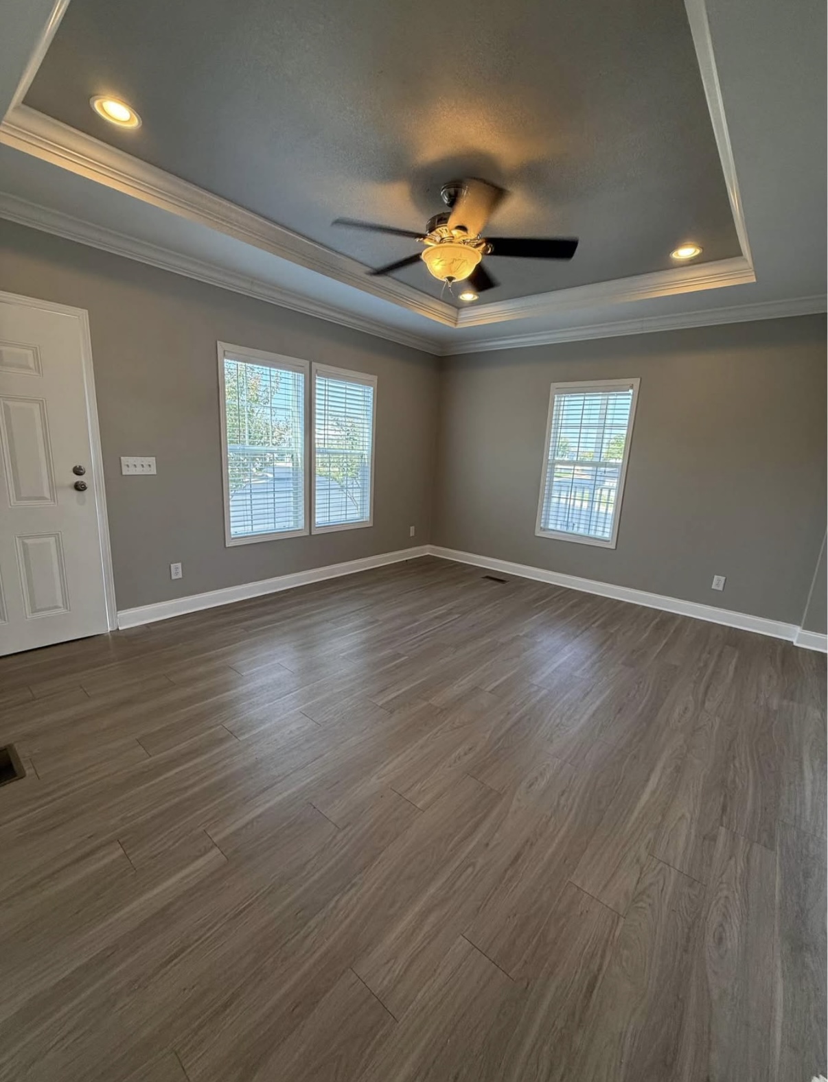 Empty living room with light wood flooring, gray walls, three windows, ceiling fan with light, and tray ceiling detail.