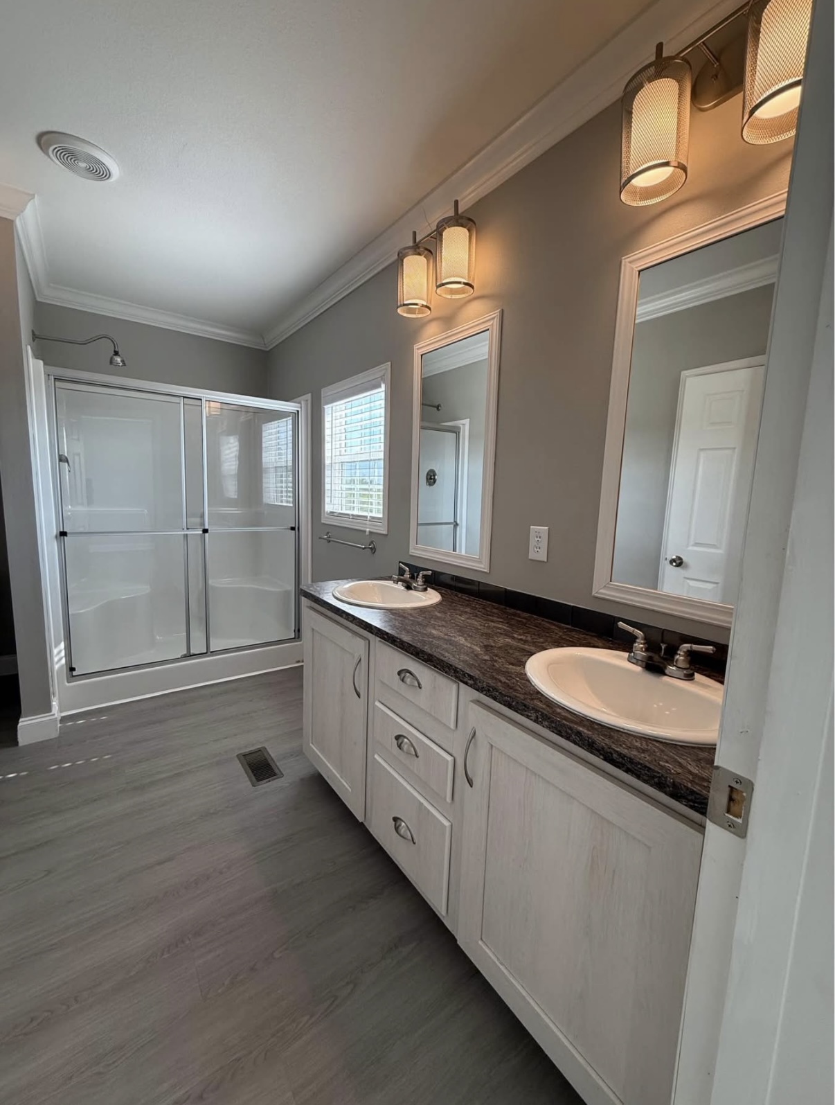 Modern bathroom with double sink vanity, dark countertop, two mirrors, overhead lights, and a glass-enclosed shower with sliding doors. Light gray walls and wood-look flooring.