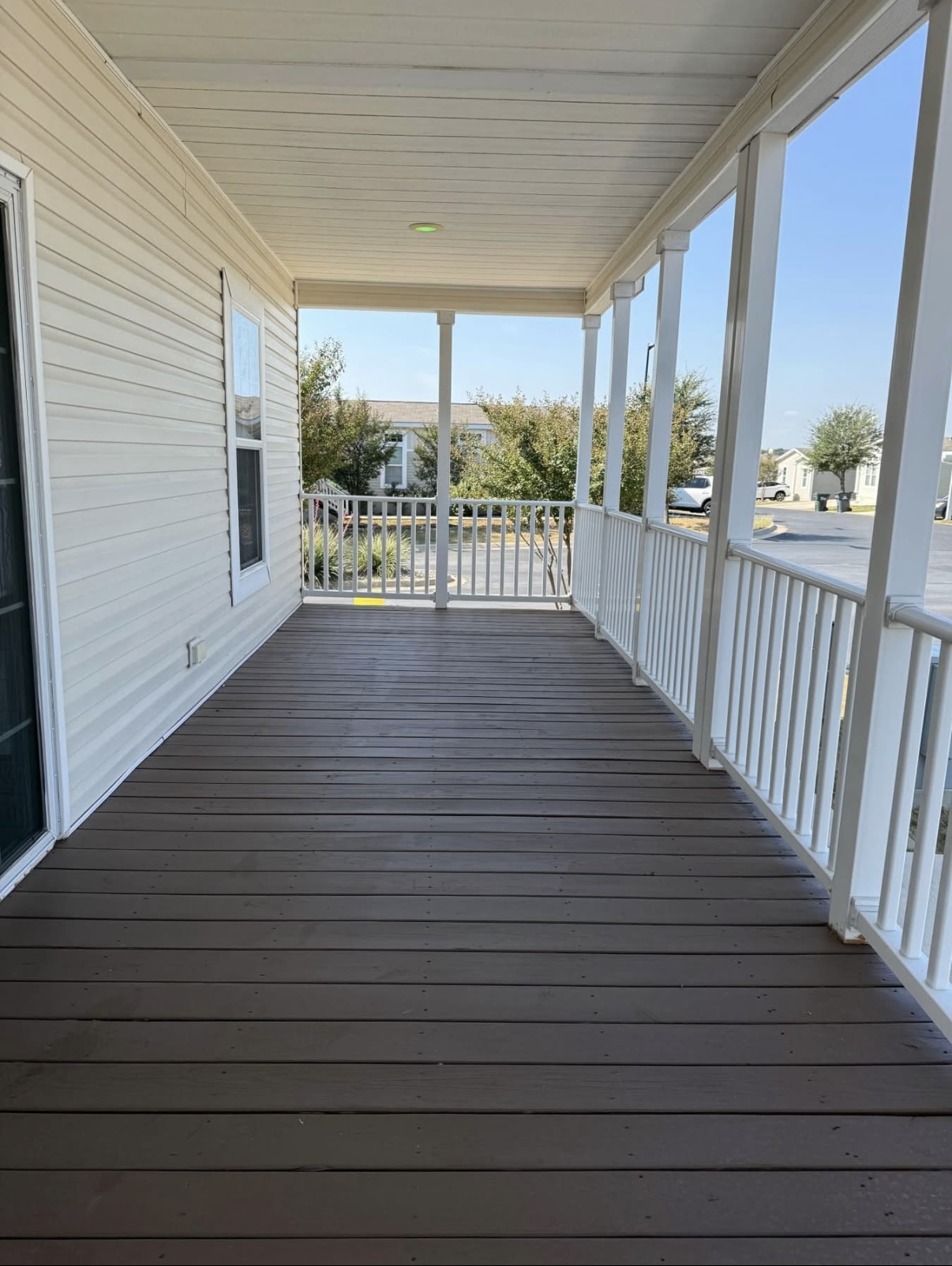 A covered porch with brown wooden flooring, white railings, and white siding, overlooking a street and trees in a residential neighborhood.