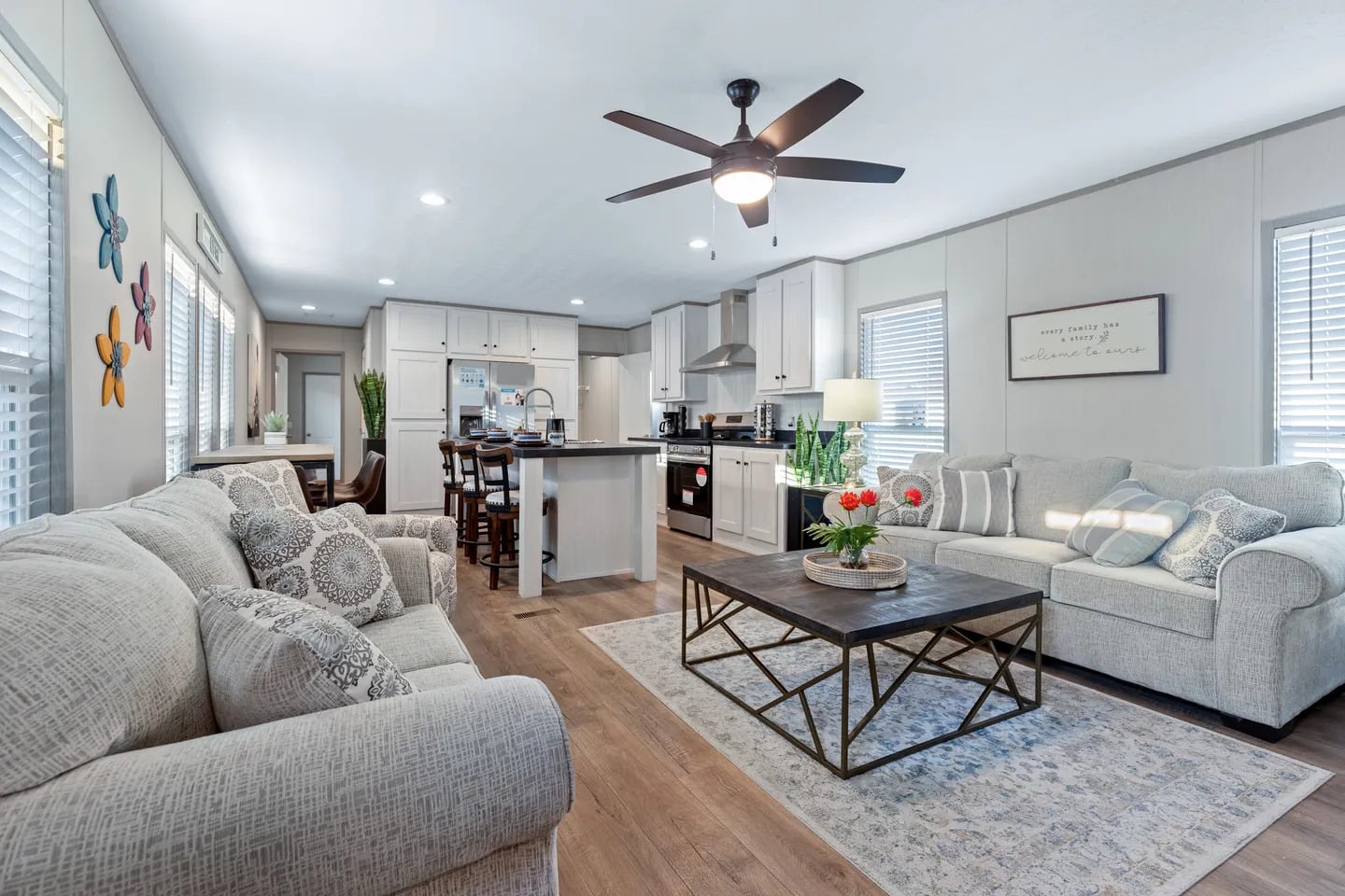 Modern open-concept living room and kitchen with neutral-toned furniture, a ceiling fan, wall art, and a central coffee table with flowers.