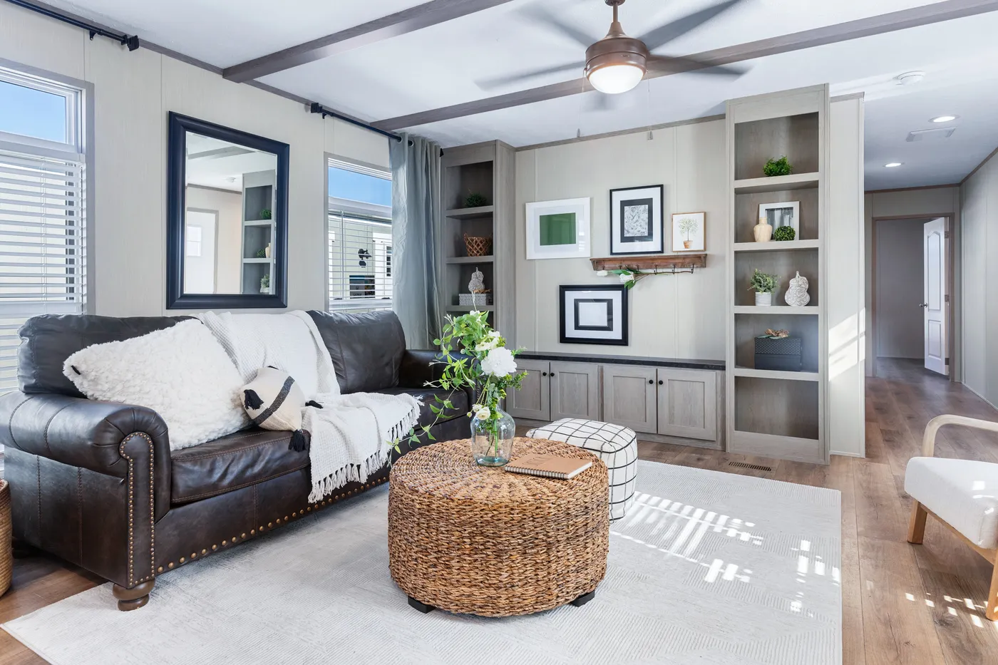 Modern living room with a dark leather sofa, woven ottoman coffee table, built-in shelves with decor, light wood floors, and neutral colors. Ceiling fan and natural light from windows.