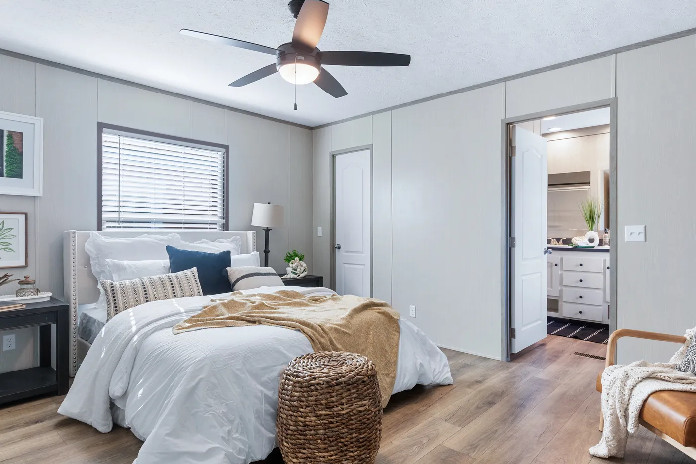 A modern bedroom with a neatly made bed, a ceiling fan, a window with blinds, light wood flooring, and an open door leading to a bathroom.