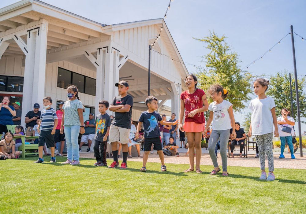 A group of children stand in a line on grass outdoors, with adults watching from behind in front of a white building under a sunny sky.